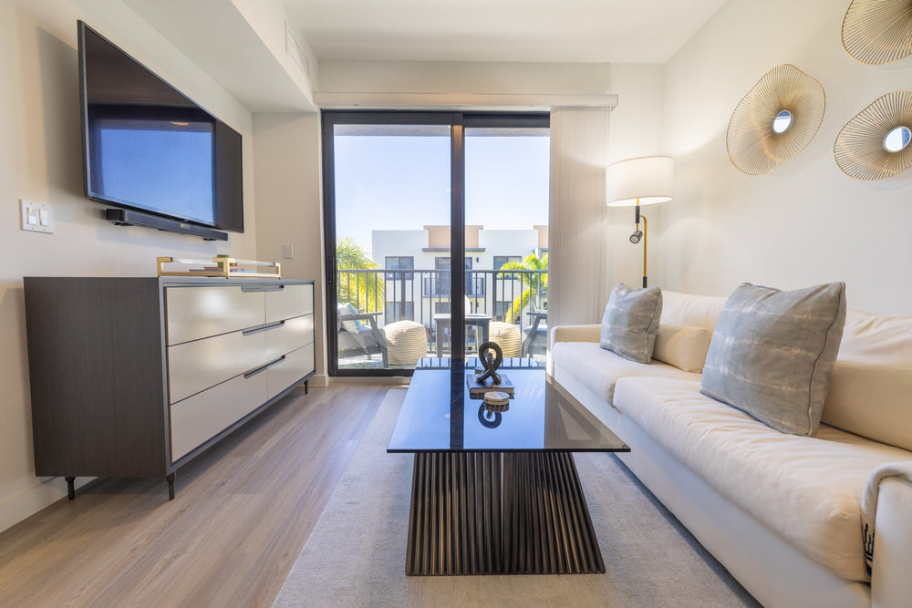 Completed apartment living area with a gray dresser, cream sofa, glass coffee table and balcony doors.