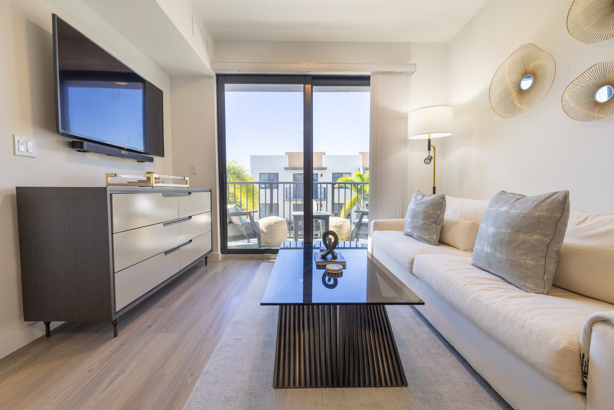 Completed apartment living area with a gray dresser, cream sofa, glass coffee table and balcony doors.